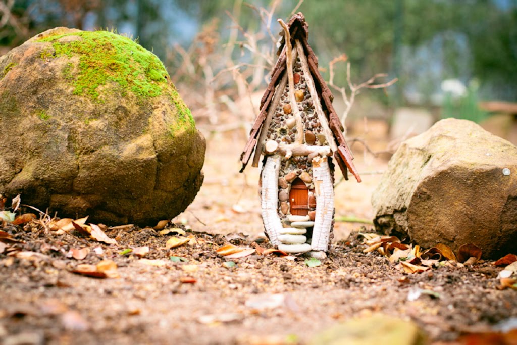 A small fairy house sits in between two large boulders, one is covered in green moss. The house has a steep, brown roof.  The red front door has five stone steps that lead up to it. 