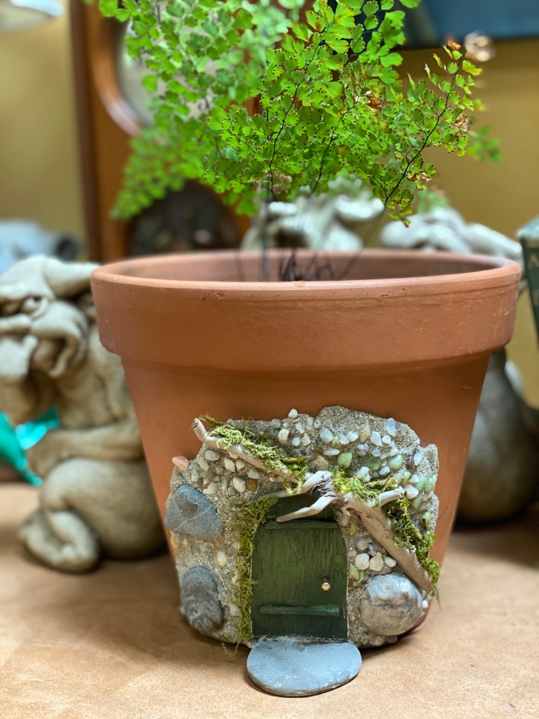 A ten inch wide terra cotta pot with a fairy door and stones on one side of it. The door is green, and a twig reaches out above the door. A maidenhair fern is planted inside the pot. 