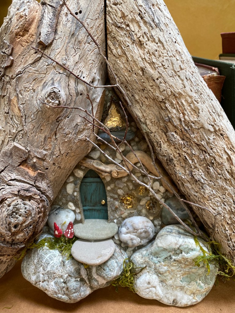 A fairy portal (a wall hanging of a single fairy  house wall).  Large wooden A-frame structure with tiny green door and green and white stone foundation.  Tiny red and white mushrooms near the door.